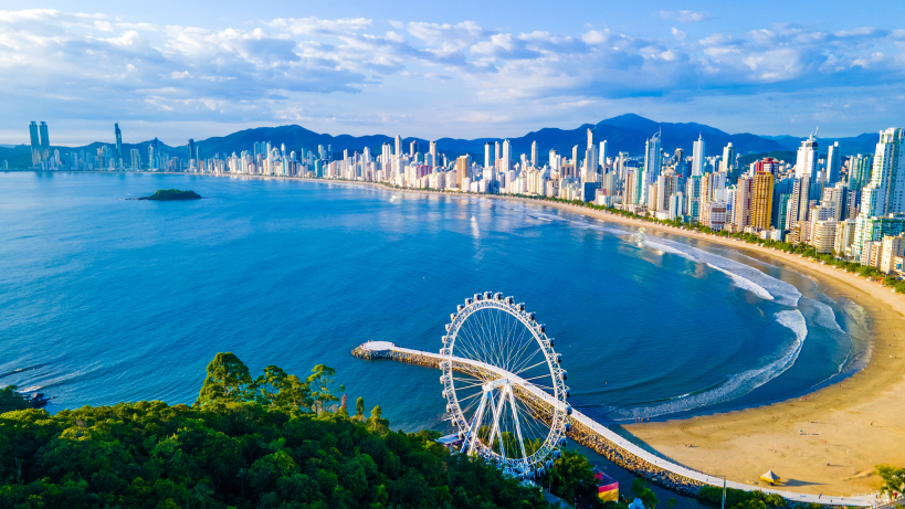 Vista panorâmica da orla de Balneário Camboriú com seus arranha-céus e a roda-gigante BC Big Wheel ao fundo, à beira da praia.