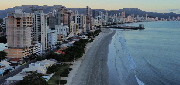 Vista aérea da Praia de Balneário Perequê em Porto Belo, Santa Catarina, com mar azul, faixa de areia clara e edifícios à beira-mar.