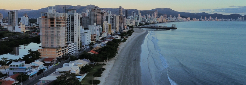 Vista aérea da Praia de Balneário Perequê em Porto Belo, Santa Catarina, com mar azul, faixa de areia clara e edifícios à beira-mar.