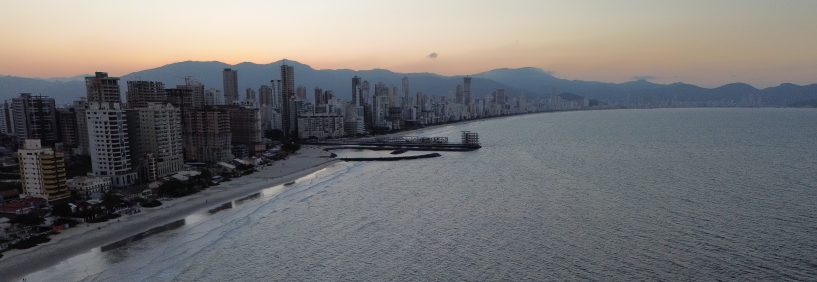 Vista aérea da Praia de Balneário Perequê em Porto Belo, Santa Catarina, com mar azul, faixa de areia clara e prédios à beira-mar.