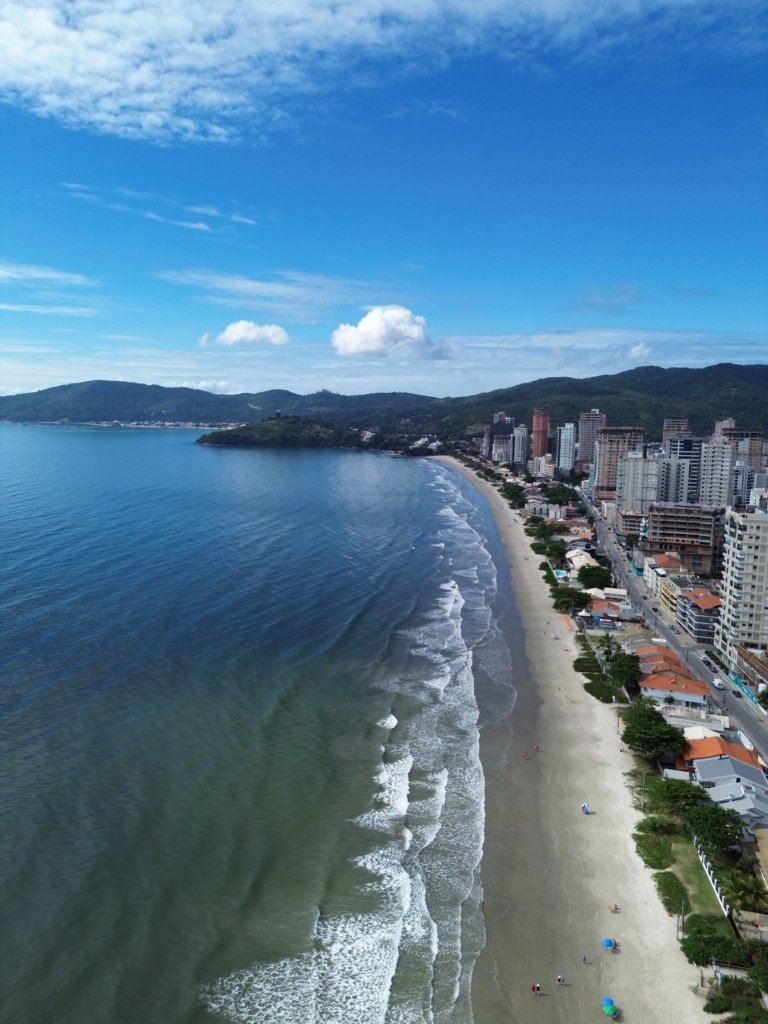 Vista aérea de drone mostrando prédios à beira-mar em Porto Belo, com a praia de Balneário perequê, areia clara e o mar azul ao fundo.